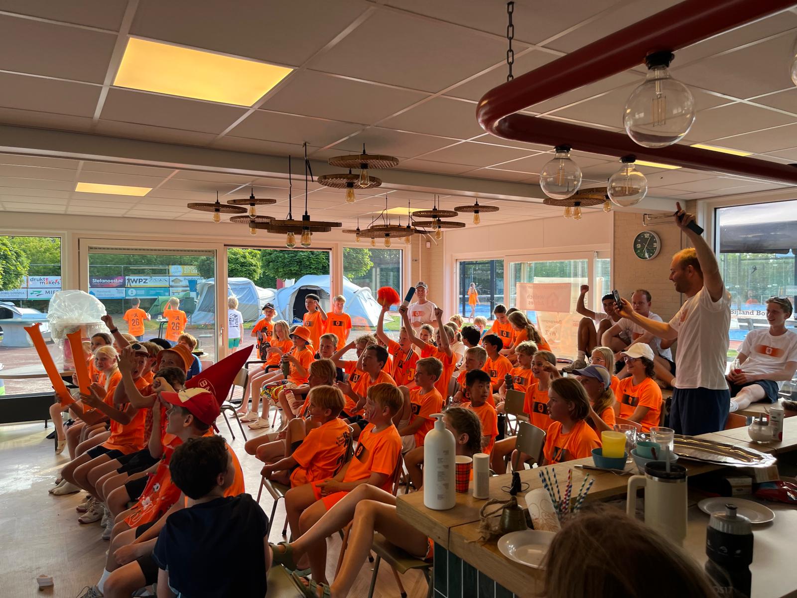 Jeugddeelnemers in oranje shirts juichen in het clubhuis tijdens het tentenkamp